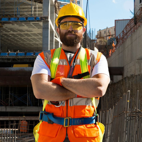 A bearded man dressed in high-visibility gear stands facing us with his arms crossed, while looking off-screen. He appears to be in a construction site.