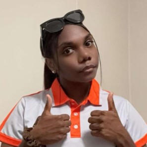 Geneva—a young First Nations woman—stands facing the camera and giving double-thumbs up, wearing a white and orange polo shirt