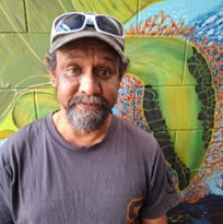 Michael—a mature-aged Torres Strait Islander man—stands facing the camera in front of an illustrated green wall, wearing a baseball cap with a grey t-shirt