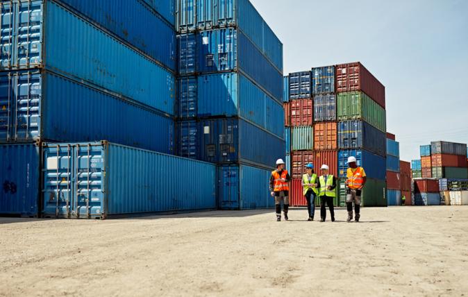 From a distance we see four people with yellow and orange vests walking together through a shipping container storage area.