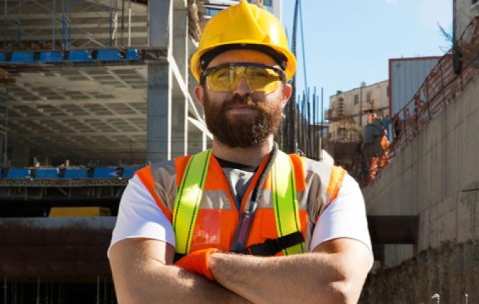 A bearded man dressed in high-visibility gear stands facing us with his arms crossed, while looking off-screen. He appears to be in a construction site.