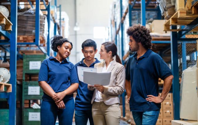 In a warehouse with blue shelves, three 20-something workers in deep blue shirts are gathered around a 30-something woman in a beige suit who is showing them something on the laptop she holds.