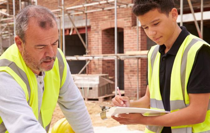 Two men dressed in high visibility vests are looking down on a table, examining documents. The men are located outside in a building site.