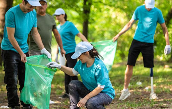 A team of 5 people in an open space environment surrounded by trees wearing gardening gloves picking up garden waste and placing in garden waste bags