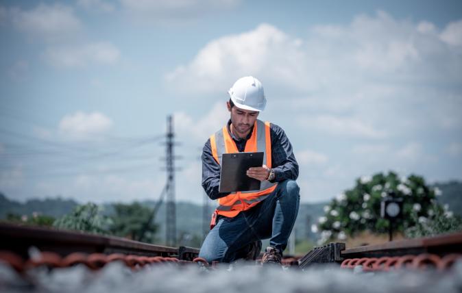 Railway worker wearing high-visibility clothing and a hard hat, kneeling down in the centre of the rail line holding a clipboard writing notes