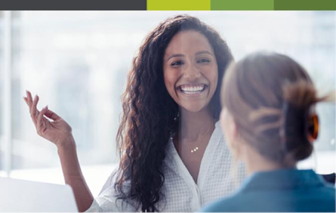 Two women in their 20s are having a joyful conversation in an office environment.