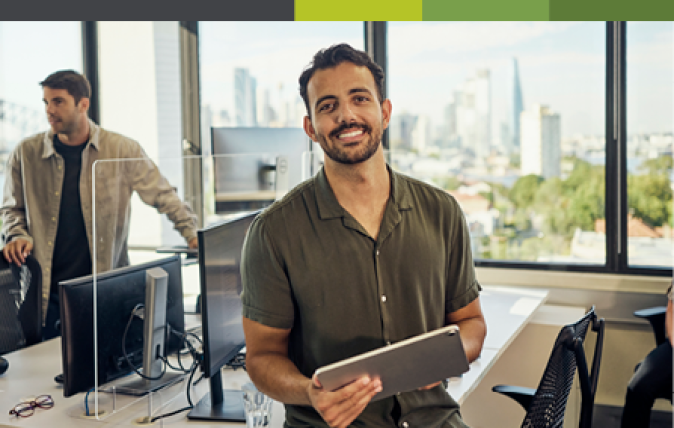 Mid shot of a smiling young man holding a tablet and standing next to a workstation in an open office.